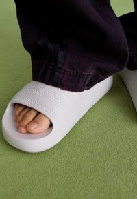 White slide sandals with textured straps and a smooth, rounded sole. Worn with dark jeans against a green background.