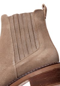 Bottine en suède beige avec des accents de couture verticaux, bout rond et un talon en bloc de bois. Texture lisse et design minimaliste.