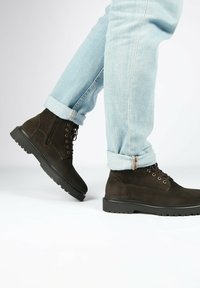 Stivaletti alla caviglia in suede marrone con suola in gomma nera, caratterizzati da sei asole e design con lacci. Indossati con jeans azzurri a risvolto.