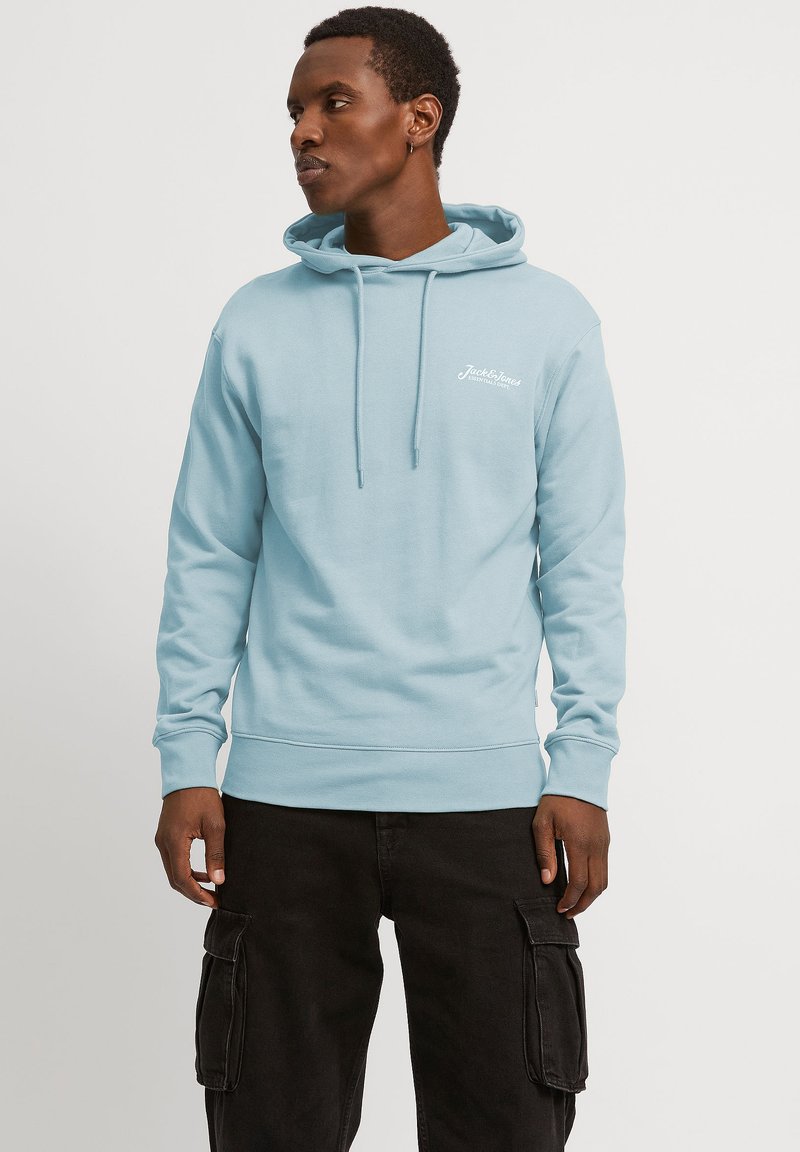 Hellblauer Hoodie aus weichem Stoff, mit einer vorderen Kängurutasche, Zugbändern und kleinem weißen Logo auf der linken Brust. Kombiniert mit schwarzen Cargo-Hosen.
