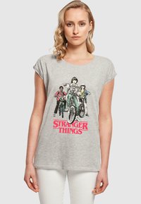 Hallhalli puuvillane T-särk, millel on graafika kolmest tegelasest jalgrattaga, allpool disaini on suurte punaste tähtedega "STRANGER THINGS".