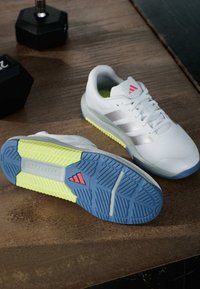 Białe buty sportowe z niebiesko-żółtymi podeszwami, czerwonym logo na podeszwie i języku, stojące na drewnianej podłodze obok czarnych hantli.