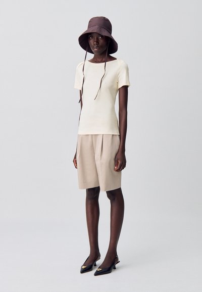 Top corto a maniche corte di color crema chiaro, pantaloni beige fino al ginocchio e cappello a secchiello marrone scuro. Tacchi a punta neri con accenti dorati.