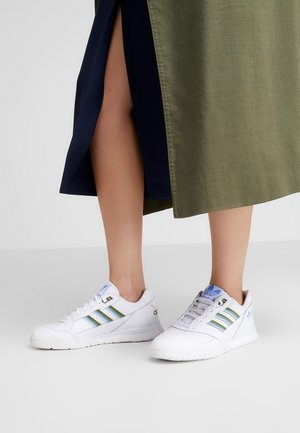 Personne portant des baskets blanches Adidas avec des rayures bleues et vertes, associées à une jupe bicolore avec une fente à l'avant.