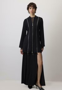 Crna haljina s dugim rukavima, prednjim zatvaračem i srebrnim detaljima. Ima prorez do buta i lepršavi kroj. Glatka tekstura tkanine.