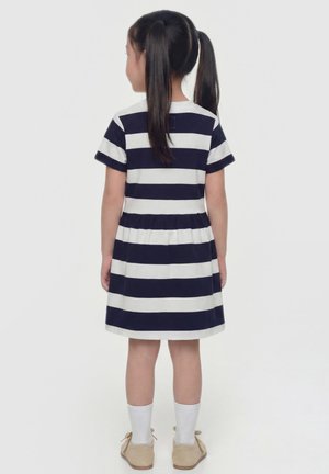 Kind met zwart haar in vlechten, gekleed in een marineblauw-wit gestreepte jurk, witte sokken en beige schoenen, staand met de rug naar voren op een witte achtergrond.