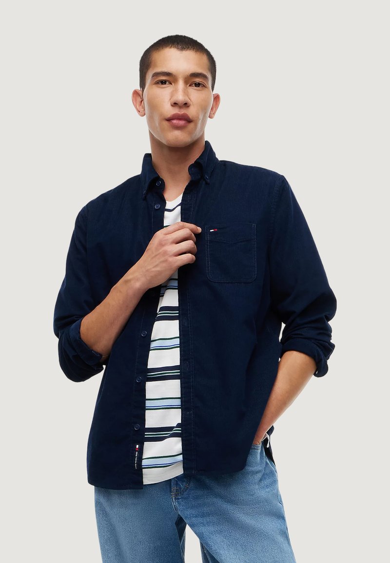 Man wearing navy button-up shirt over white striped t-shirt and light blue jeans, with one hand adjusting shirt and the other in pocket.