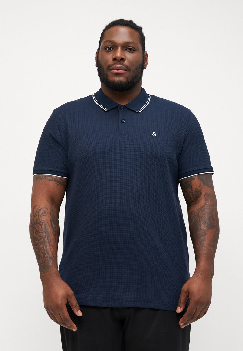 Tricou polo de culoare bleumarin, dintr-un material texturat, cu o bandă albă pe guler și mâneci, și un mic logo pe piept.