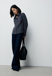 Navy gestreept overhemd met knopen, donkerblauwe denim wijde broek, zwarte handtas met een gladde textuur, zwarte spitse hakken, neutrale achtergrond.