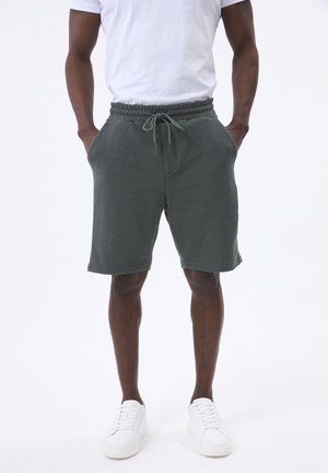 Groene gebreide short voor heren met een elastische tailleband en een trekkoord. Voorzien van zijzakken en een textuurpatroon. Gecombineerd met witte sneakers.