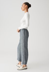 Witte lange mouwen top, lichtblauwe linnen broek met trekkoord en beige instap schoenen, weergegeven vanuit de zijkant op een effen achtergrond.