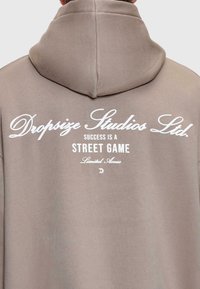 Beige Hoodie mit weißem Text auf dem Rücken, der "Dropsize Studios Ltd. Erfolg ist ein Straßen-Spiel mit eingeschränktem Zugang" liest.