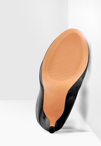 Chaussure noire vue par le dessous, avec une semelle lisse à surface texturée et un talon arrondi, sur un fond gris clair.