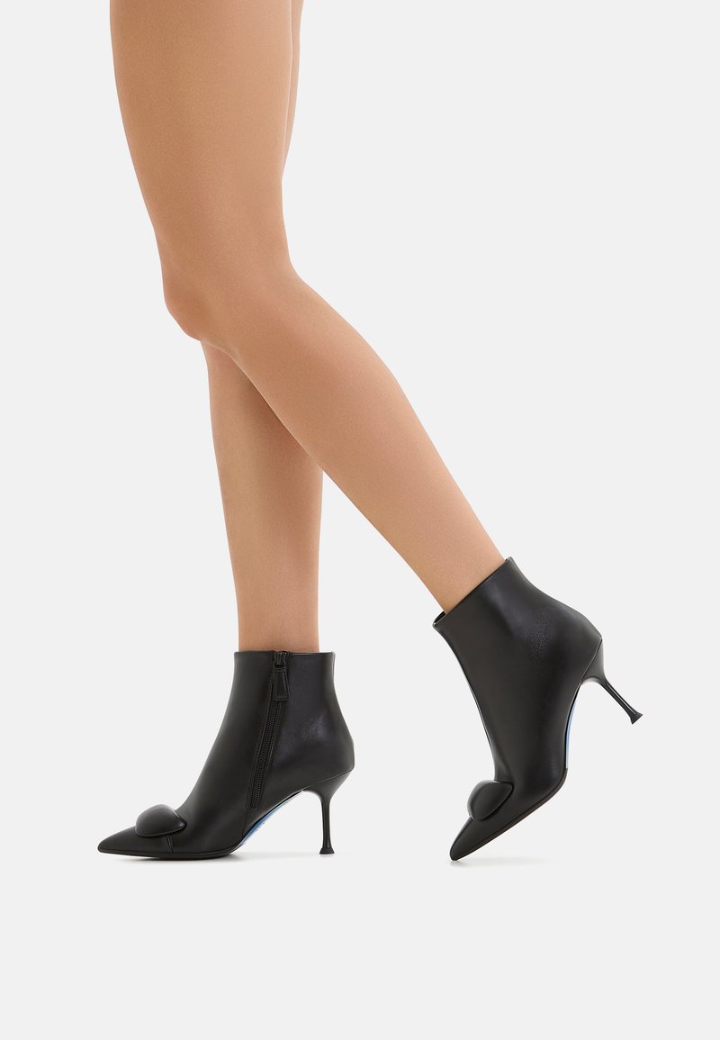 Stivali alla caviglia in pelle nera con punta affusolata, chiusura con zip e un piccolo dettaglio decorativo sulla punta. Presentano un tacco alto sottile.