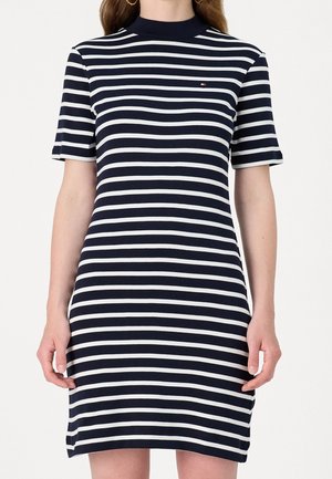 Vrouw die een kort mouwjurk draagt met horizontale marineblauwe en witte strepen, met een klein logo op de borst en gouden oorringen.