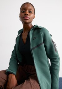 Zelená Nike mikina na zips s priliehavým strihom, čiernymi akcentmi a vreckom na zips. Nosená cez čierny top, v kombinácii s hnedými nohavicami.