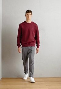 Sudadera burdeos, con puños de canalé, combinada con pantalones a rayas grises; el modelo lleva zapatillas blancas. Ambiente minimalista con suelo de madera.