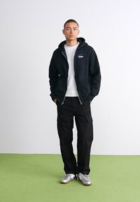 Ung mand står med hænderne i lommerne iført en sort zip-up hættetrøje, hvid skjorte, sorte cargobukser og sølvfarvede sneakers på grøn gulv.