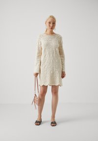 Robe en crochet crème à manches longues et ourlet en zigzag, présentant des motifs circulaires. Associée à un sac à main rose et des sandales noires.