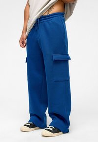 Blauwe joggers van zacht materiaal, met een comfortabele pasvorm, een elastische taille met trekkoord en twee zijcargozakken. Gecombineerd met zwarte sneakers.