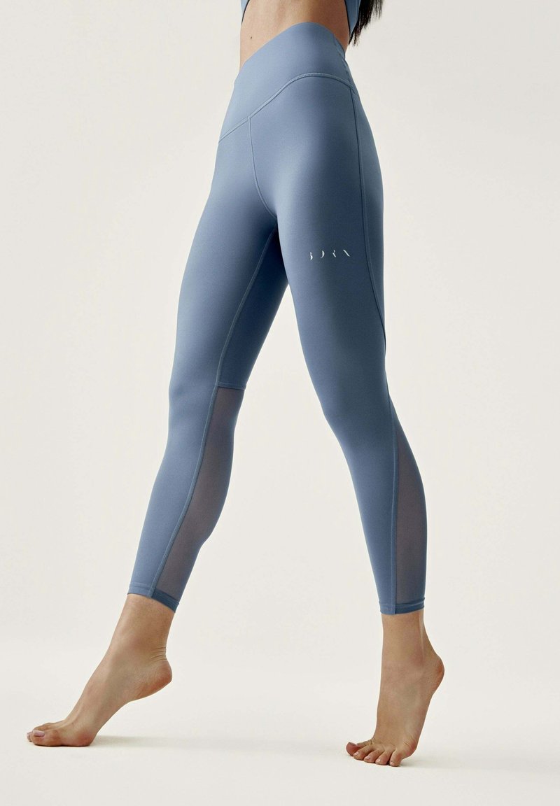 Kvinna i högmidjade blå leggings med nätpaneler på vadarna, stående barfota med ena foten något lyft på en neutral bakgrund.