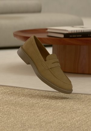 Mocassin en daim marron flottant au-dessus d'un tapis beige texturé dans un salon avec une table basse en bois et des livres empilés.