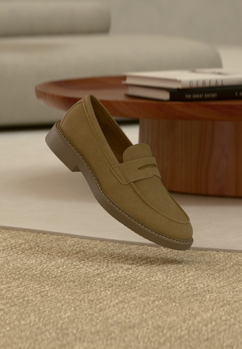 Mocassin en daim marron flottant au-dessus d'un tapis beige texturé dans un salon avec une table basse en bois et des livres empilés.