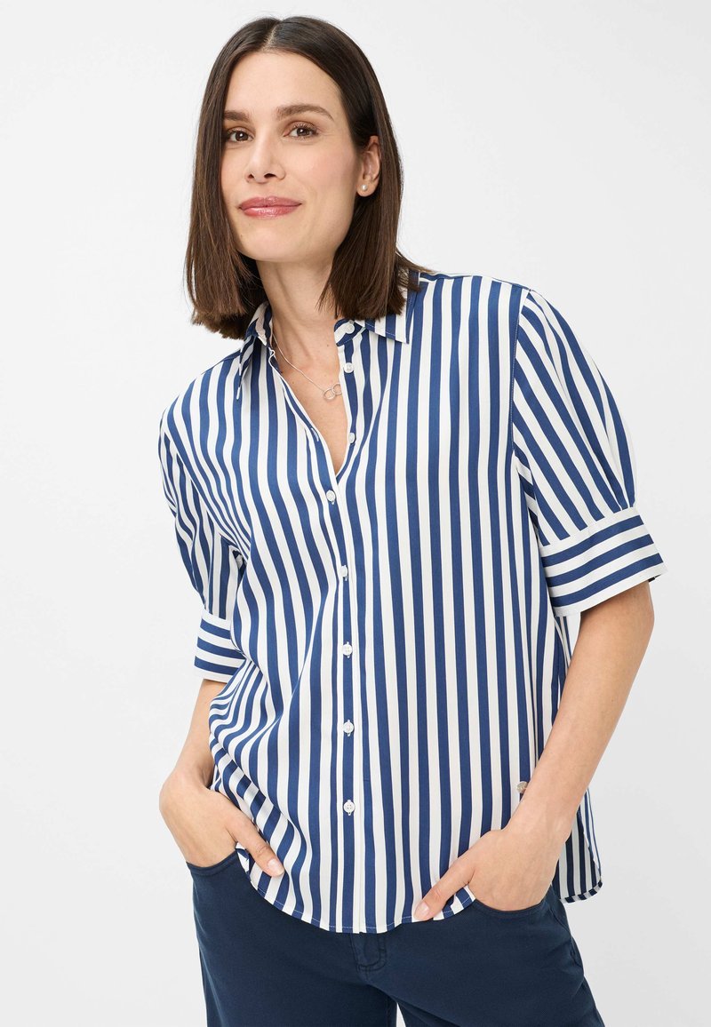 Camicia a maniche corte con bottoni, caratterizzata da righe verticali blu e bianche, realizzata in tessuto morbido. Le mani del modello sono nelle tasche, vestibilità rilassata.