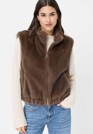 Frau trägt eine braune, ärmellose Weste aus Kunstfell über einem cremefarbenen Pullover und blauen Jeans, steht mit einer Hand in der Tasche.