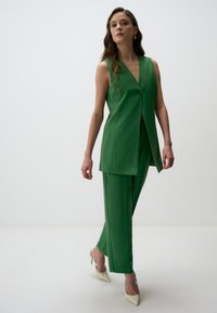 Gilet vert ajusté avec un profond décolleté en V et un bouton unique, associé à un pantalon large assorti et des escarpins crèmes à bout pointu.
