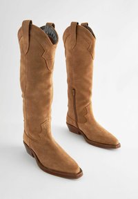 Magas barna velúr cowboy csizmák hegyes orral, díszes varrással és oldalsó cipzárokkal. Felsőrészén húzófülek találhatóak a könnyű felvétel érdekében.