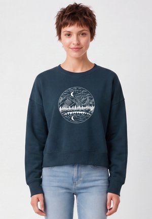 Junge Frau mit kurzen braunen Haaren trägt einen dunkelblauen Pullover mit weißem, rundem Berg- und Baummotiv, kombiniert mit hellblauen Jeans.