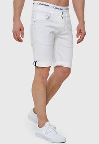 Shorts en denim blanc avec des revers, présentant un accent bleu sur le revers, et une taille de marque Calvin Klein. Portés avec des baskets blanches.