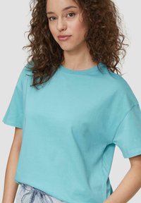 Turkusowa bawełniana koszulka z okrągłym dekoltem, krótkimi rękawami i luźnym krojem, prezentowana na neutralnym tle.