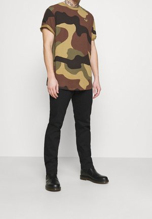 T-shirt à motif camouflage dans des nuances de marron et de vert, associé à un pantalon slim noir et des bottines en cuir noires. Esthétique décontractée.
