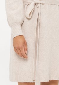 Brede gebreide jurk in zacht beige met geribde manchetten en een strikceintuur. Heeft een ontspannen pasvorm en een textielstructuur voor warmte en comfort.