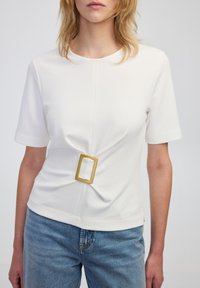 Weißes kurzärmliges Oberteil mit einer verdrehten Vorderseite und einem rechteckigen goldfarbenen Hardware-Akzent an der Taille. Glattes Material und runder Ausschnitt.