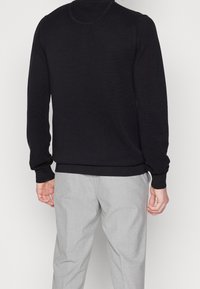 Homme portant un pull noir texturé à manches longues et un pantalon gris clair, debout de dos face à la caméra sur un fond blanc.