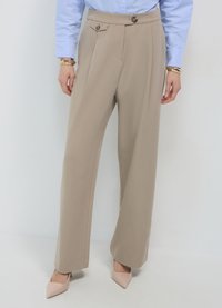 Beige maßgeschneiderte Hose mit hoher Taille, ausgestattet mit doppelten Knöpfen und Falten, kombiniert mit hellrosa spitzen High Heels. Glatte Stofftextur.