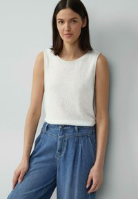 Haut sans manches blanc avec un design en maille texturée, associé à un pantalon en denim bleu taille haute présentant des plis et une coupe droite.