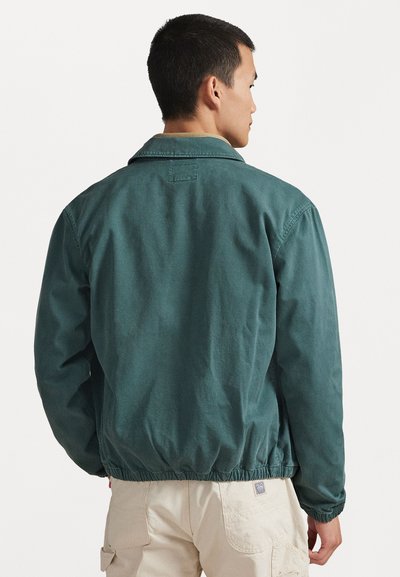 Žalia medvilninė striukė laisvo kirpimo, su nuleidžiamu apykakliu ir elastiniu liemens diržu, pasižyminti lygiu audiniu ir minimaliais siuvimo detalėmis.