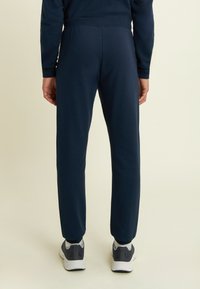 Persona che indossa pantaloni tuta blu navy con un maglione coordinato a maniche lunghe e scarpe da ginnastica grigio-bianco, in piedi su uno sfondo chiaro.