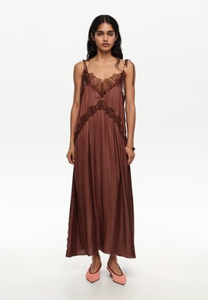 Mujer con un vestido largo marrón sin mangas con detalles de encaje y tirantes finos en los hombros, combinado con zapatos planos rosas de punta afilada.