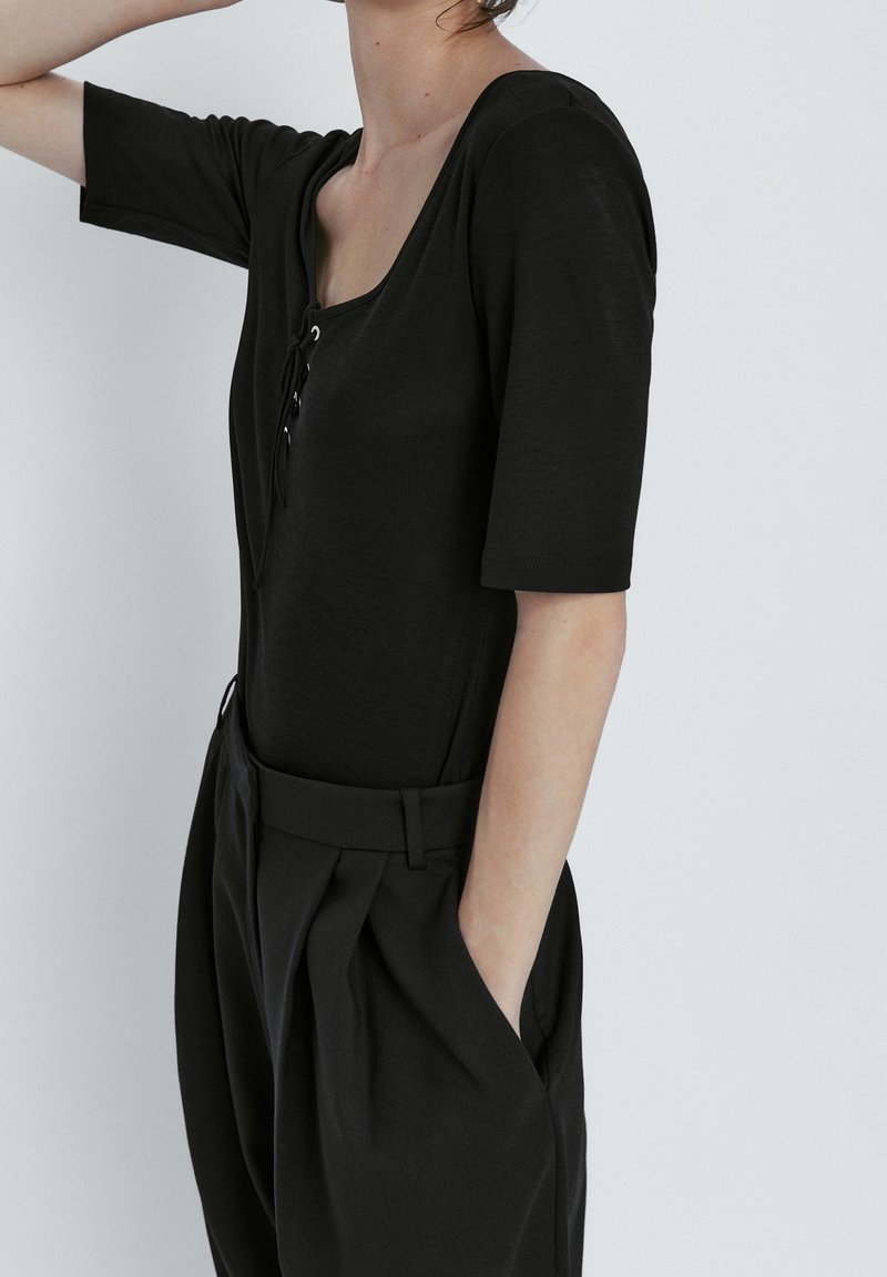 Femme portant un haut noir à manches courtes avec un col lacé et un pantalon noir taille haute, les mains dans les poches, sur un fond uni.