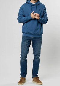Sudadera azul con cordón ajustable, puños y dobladillo acanalados, combinada con jeans azules. El modelo está de pie con las manos entrelazadas, usando zapatillas de deporte color beige.