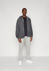 Grå hettejakke med glidelås og teksturert overflate, svart t-skjorte, lysegrå joggebukser og hvite joggesko; har hette og elastiske mansjetter.