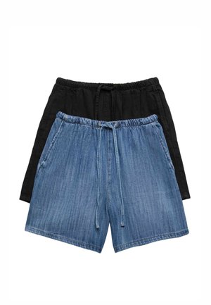 Kvinde iført solbriller, vinrød strikket cardigan, blå denimshorts og brune sandaler, stående med den ene hånd i lommen.