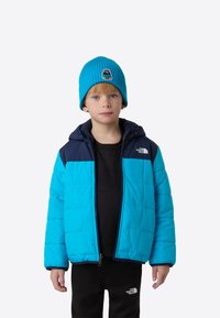 Niebieska kurtka puchowa z granatowymi akcentami, zamkiem błyskawicznym i kapturem. Do tego dopasowana prążkowana niebieska czapka z małą naszywką z misiem oraz czarne spodnie.