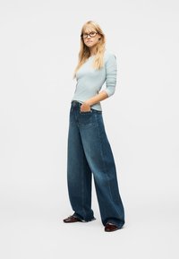 Jeune femme aux longs cheveux blonds et lunettes, portant une chemise bleu clair à manches longues, un jean large foncé et des chaussures marron, debout avec les mains dans les poches.