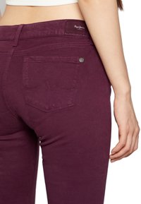 Lila skinny jeans med en jämn textur, en liten metallknapp och en bakficka med ett vågigt sömnads mönster. Märkesetikett synlig.